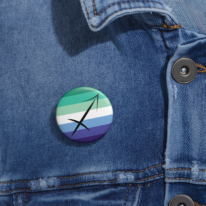 Close-up of a white circular pride button featuring the Sagittarius zodiac glyph centered over a gay men (MLM) pride flag background in greens/teals, white, and blues, attached to a blue denim jacket at the right chest with the button clearly visible.