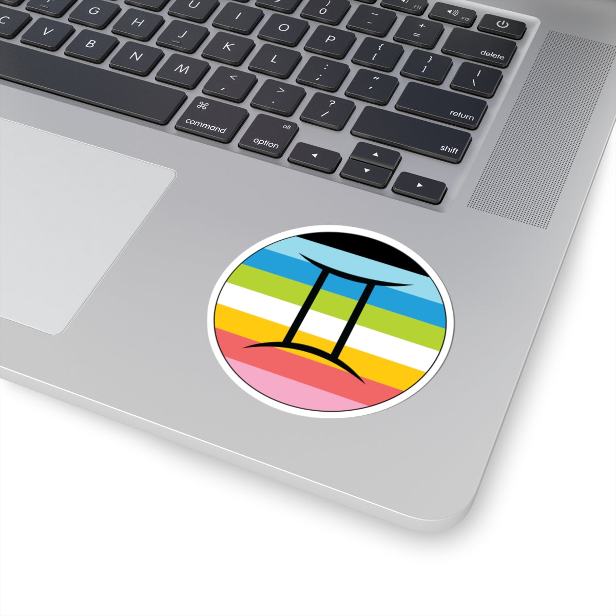 Close-up of the bottom right corner of a silver laptop featuring a 3 inch by 3 inch circular zodiac sticker with a centered Gemini glyph on a Queer pride flag style background applied to the surface.
