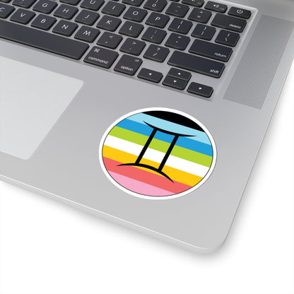 Close-up of the bottom right corner of a silver laptop featuring a 3 inch by 3 inch circular zodiac sticker with a centered Gemini glyph on a Queer pride flag style background applied to the surface.