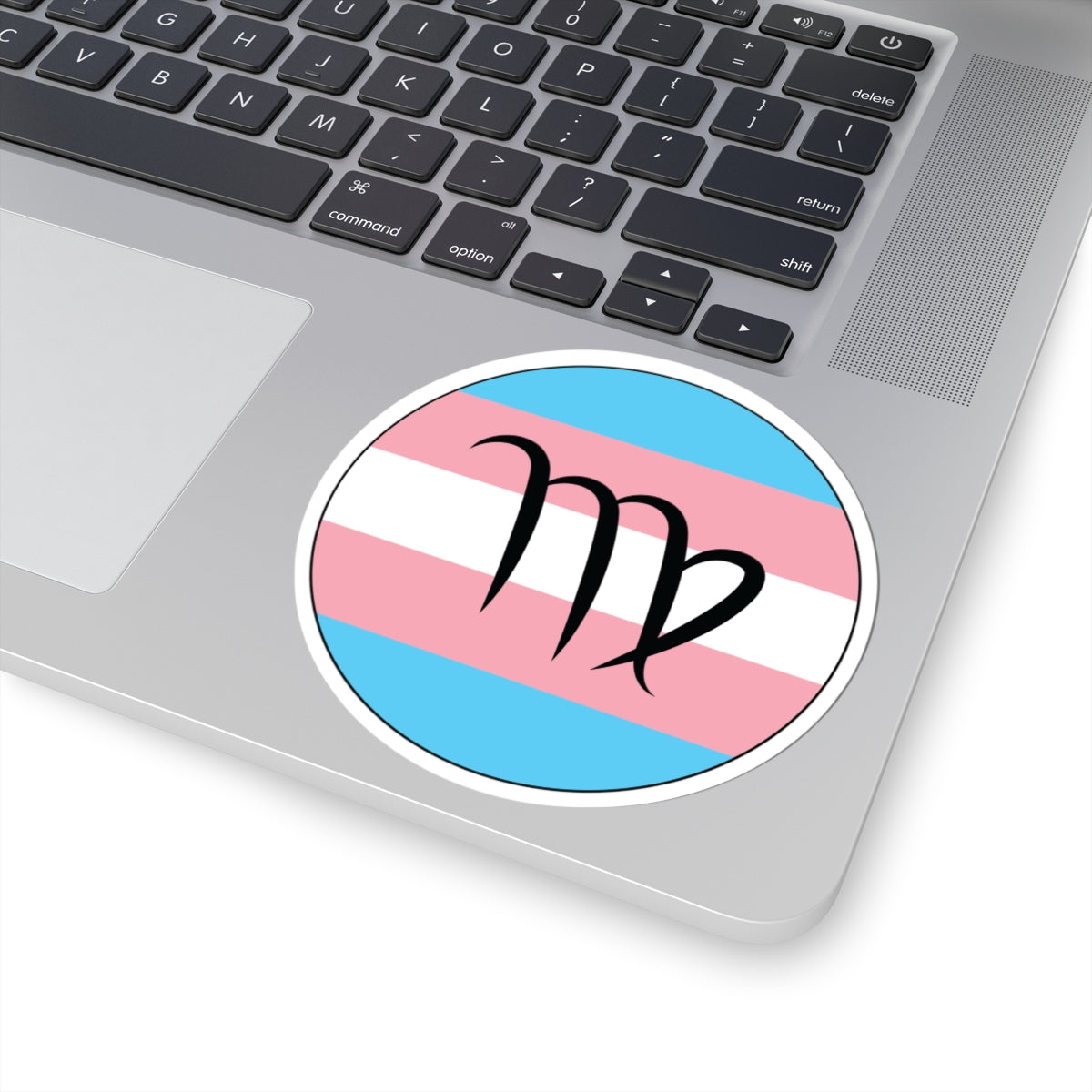 Close-up of the bottom right corner of a silver laptop featuring a 4 inch by 4 inch circular zodiac sticker with a centered Virgo glyph on a Transgender pride flag style background applied to the surface.