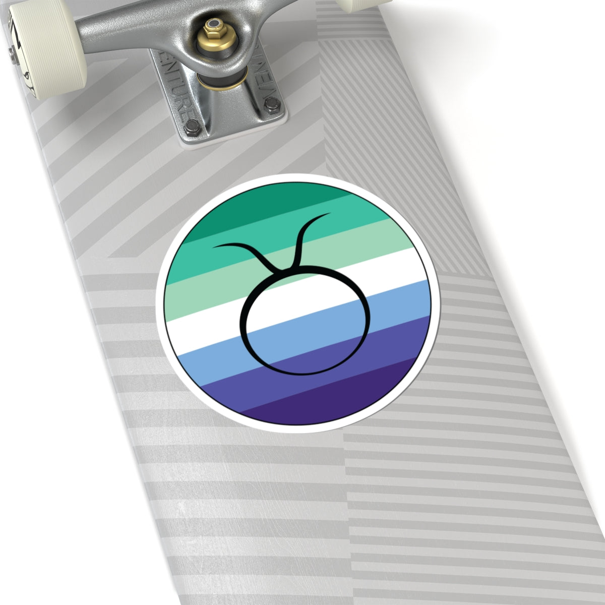 Close-up of the bottom of a silver skateboard featuring a 6 inch by 6 inch circular zodiac sticker with a centered Taurus glyph on a Gay Men pride flag style background applied to the surface.