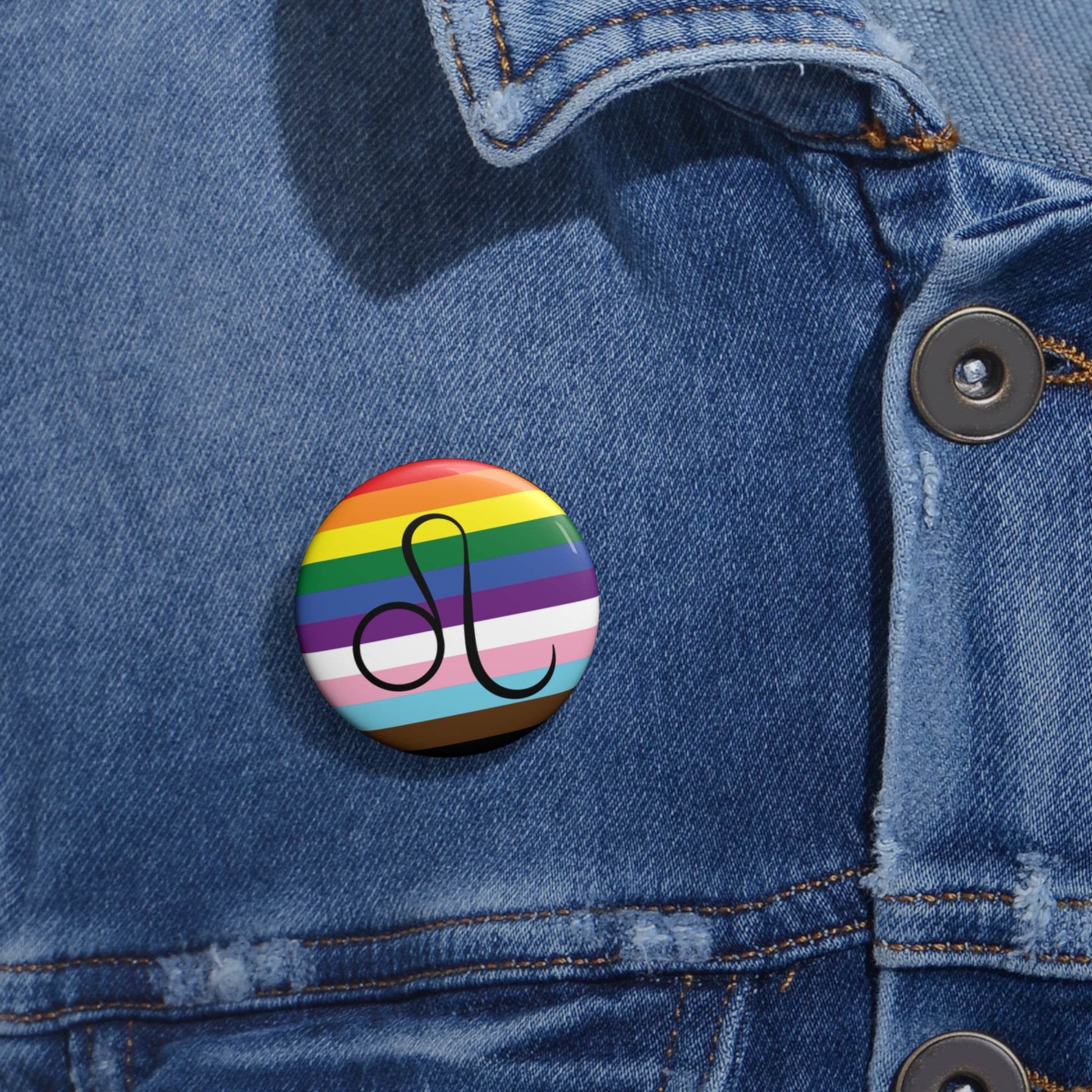 Close-up of a white circular pride button featuring the Leo zodiac glyph centered over a Progress Pride flag background in rainbow stripes with black, brown, light blue, pink, and white elements, attached to a blue denim jacket at the right chest with the button clearly visible.