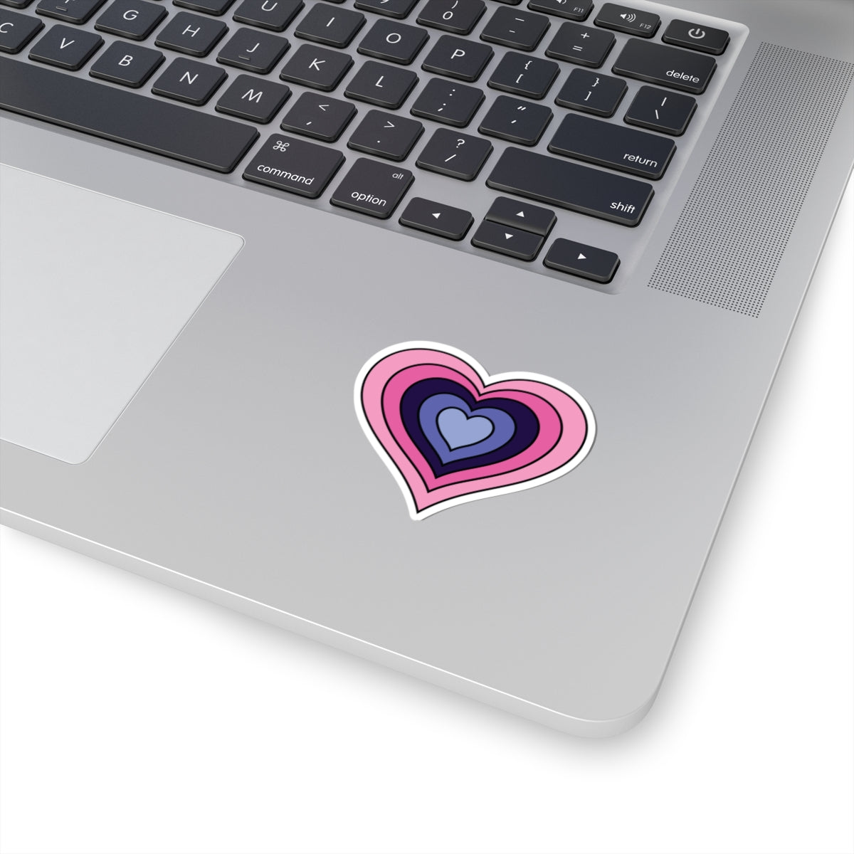 Close-up of the bottom right corner of a silver laptop featuring a 2 inch by 2 inch pride flag heart sticker in an Omnigender flag style applied to the surface.