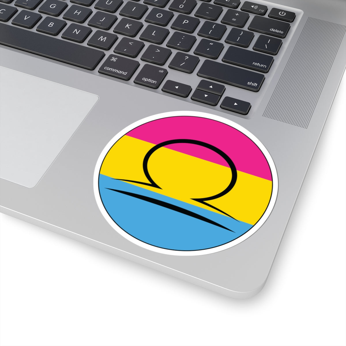 Close-up of the bottom right corner of a silver laptop featuring a 4 inch by 4 inch circular zodiac sticker with a centered Libra glyph on a Pansexual pride flag style background applied to the surface.