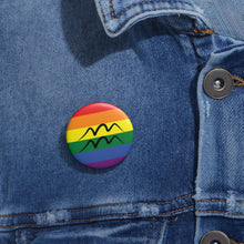 Close-up of a white circular pride button featuring the Aquarius zodiac glyph centered over a rainbow pride flag background in red, orange, yellow, green, blue, and purple, attached to a blue denim jacket at the right chest with the button clearly visible.