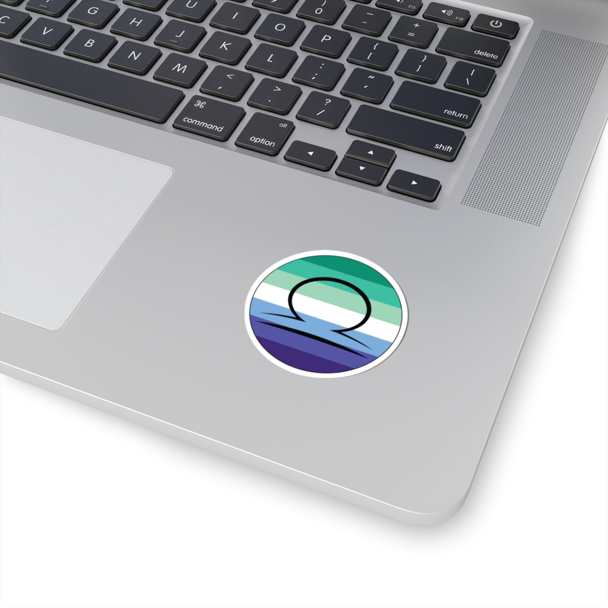 Close-up of the bottom right corner of a silver laptop featuring a 2 inch by 2 inch circular zodiac sticker with a centered Libra glyph on a Gay Men pride flag style background applied to the surface.