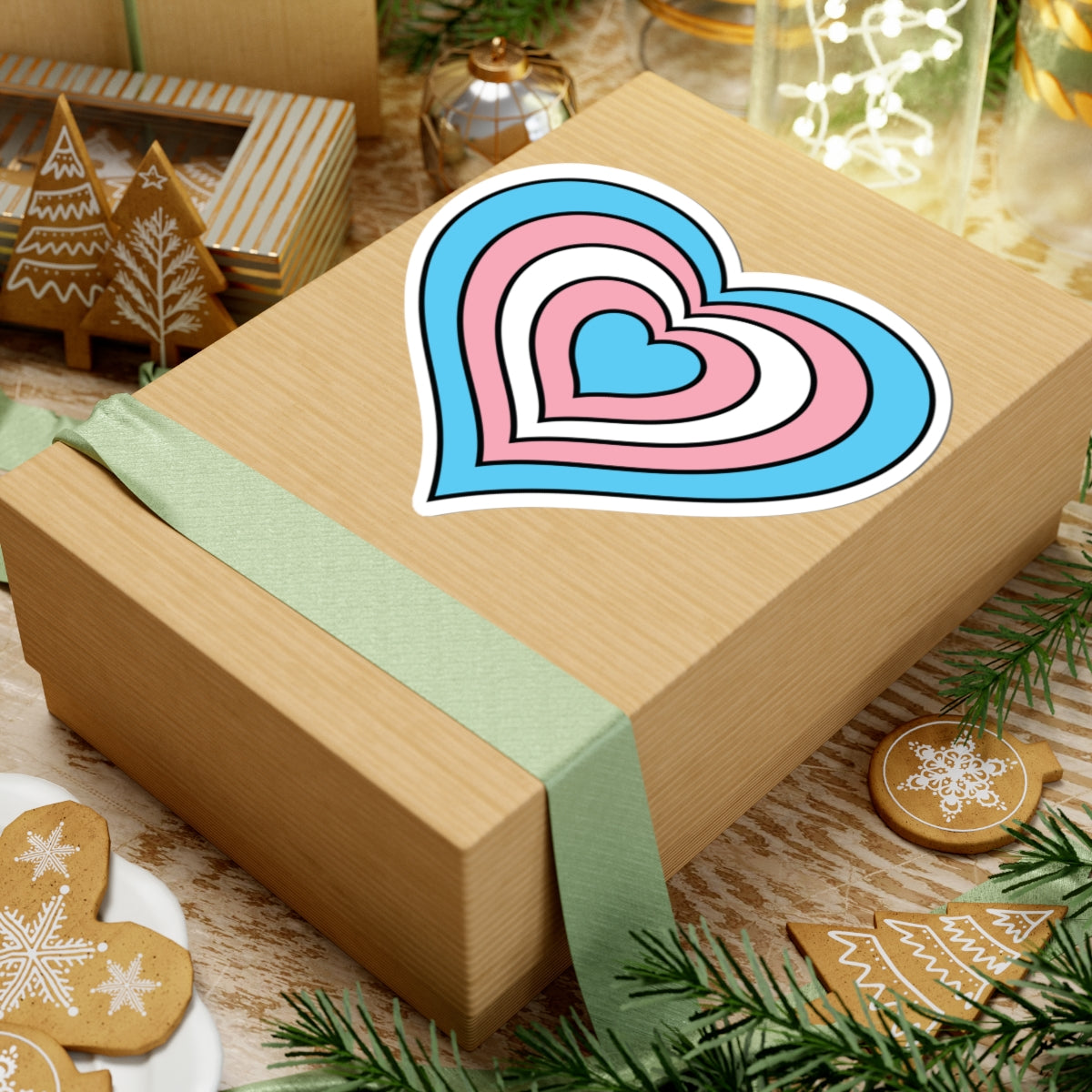 Gift box with a green ribbon on a festive background with cookies and Christmas decorations. Heart-shaped Transgender pride sticker centered on the box.