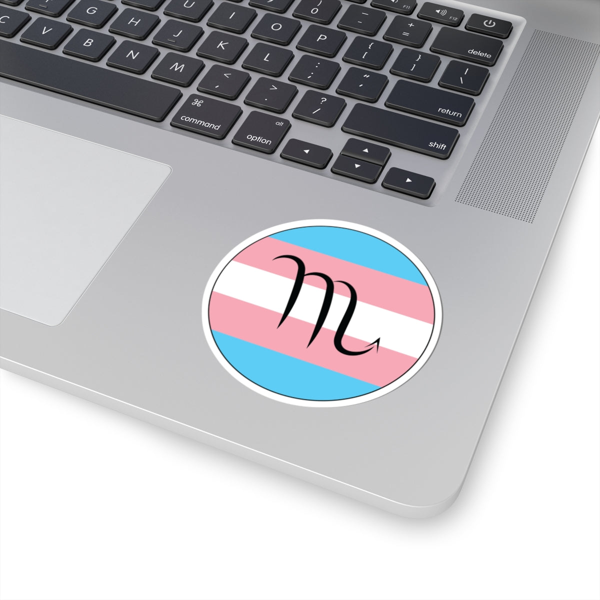 Close-up of the bottom right corner of a silver laptop featuring a 3 inch by 3 inch circular zodiac sticker with a centered Scorpio glyph on a Transgender pride flag style background applied to the surface.