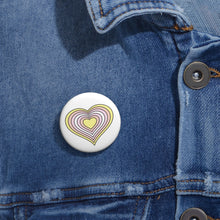 Close-up of a white circular pride button featuring a nested heart design inspired by the Pangender pride flag in yellow, oranges and white, attached to a blue denim jacket at the right chest with the button clearly visible.