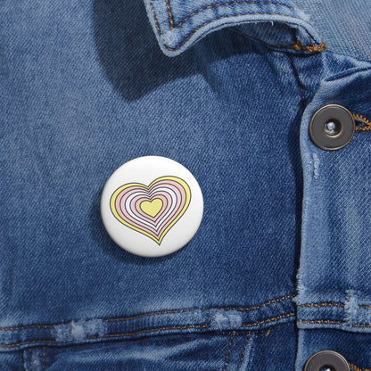 Close-up of a white circular pride button featuring a nested heart design inspired by the Pangender pride flag in yellow, oranges and white, attached to a blue denim jacket at the right chest with the button clearly visible.