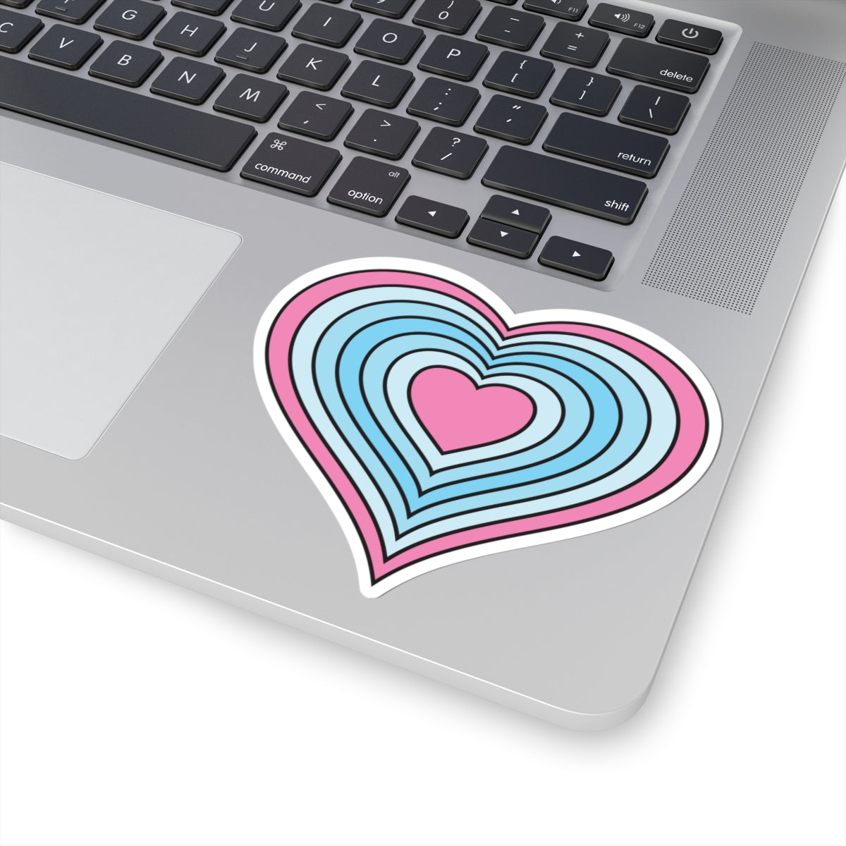 Close-up of the bottom right corner of a silver laptop featuring a 4 inch by 4 inch pride flag heart sticker in a Transmasc flag style applied to the surface.