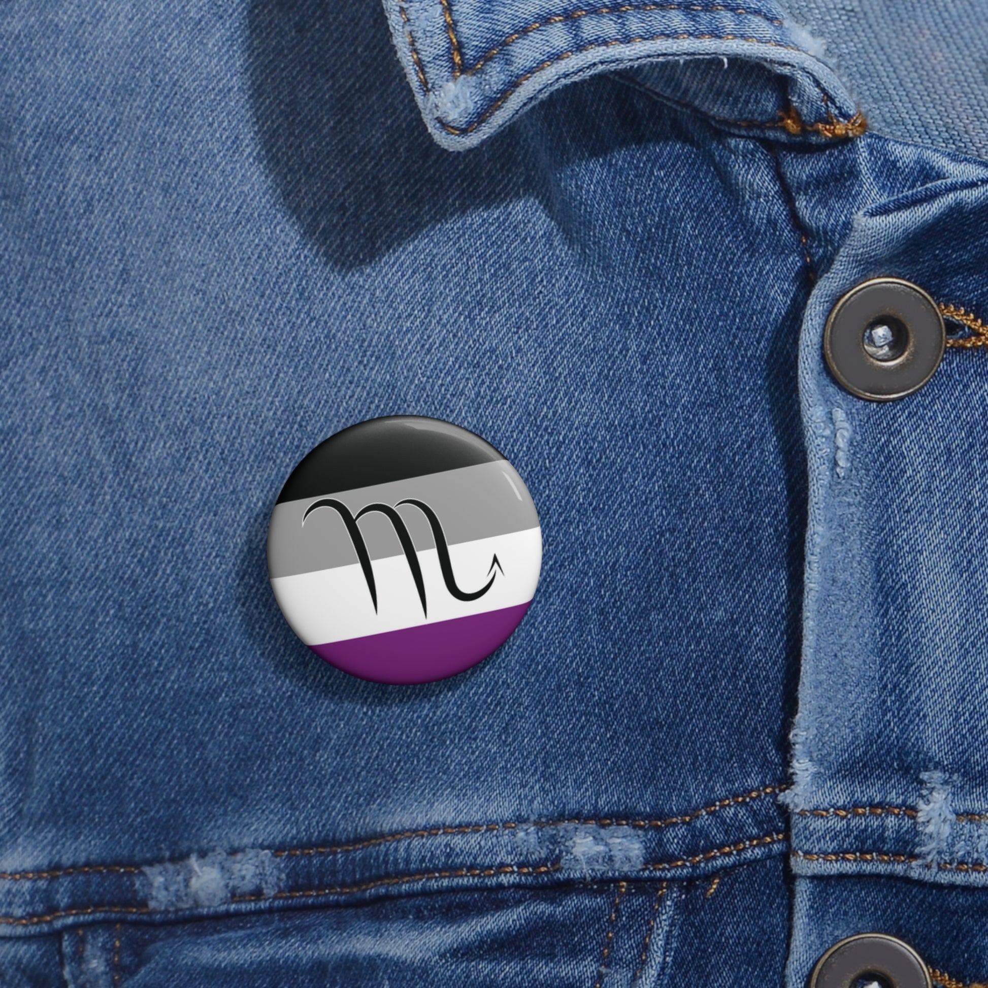 Close-up of a white circular pride button featuring the Scorpio zodiac glyph centered over an asexual pride flag background in black, gray, white, and purple, attached to a blue denim jacket at the right chest with the button clearly visible.