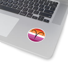 Close-up of the bottom right corner of a silver laptop featuring a 2 inch by 2 inch circular zodiac sticker with a centered Aires glyph on a Lesbian pride flag style background applied to the surface.