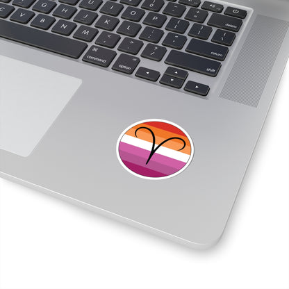 Close-up of the bottom right corner of a silver laptop featuring a 2 inch by 2 inch circular zodiac sticker with a centered Aires glyph on a Lesbian pride flag style background applied to the surface.