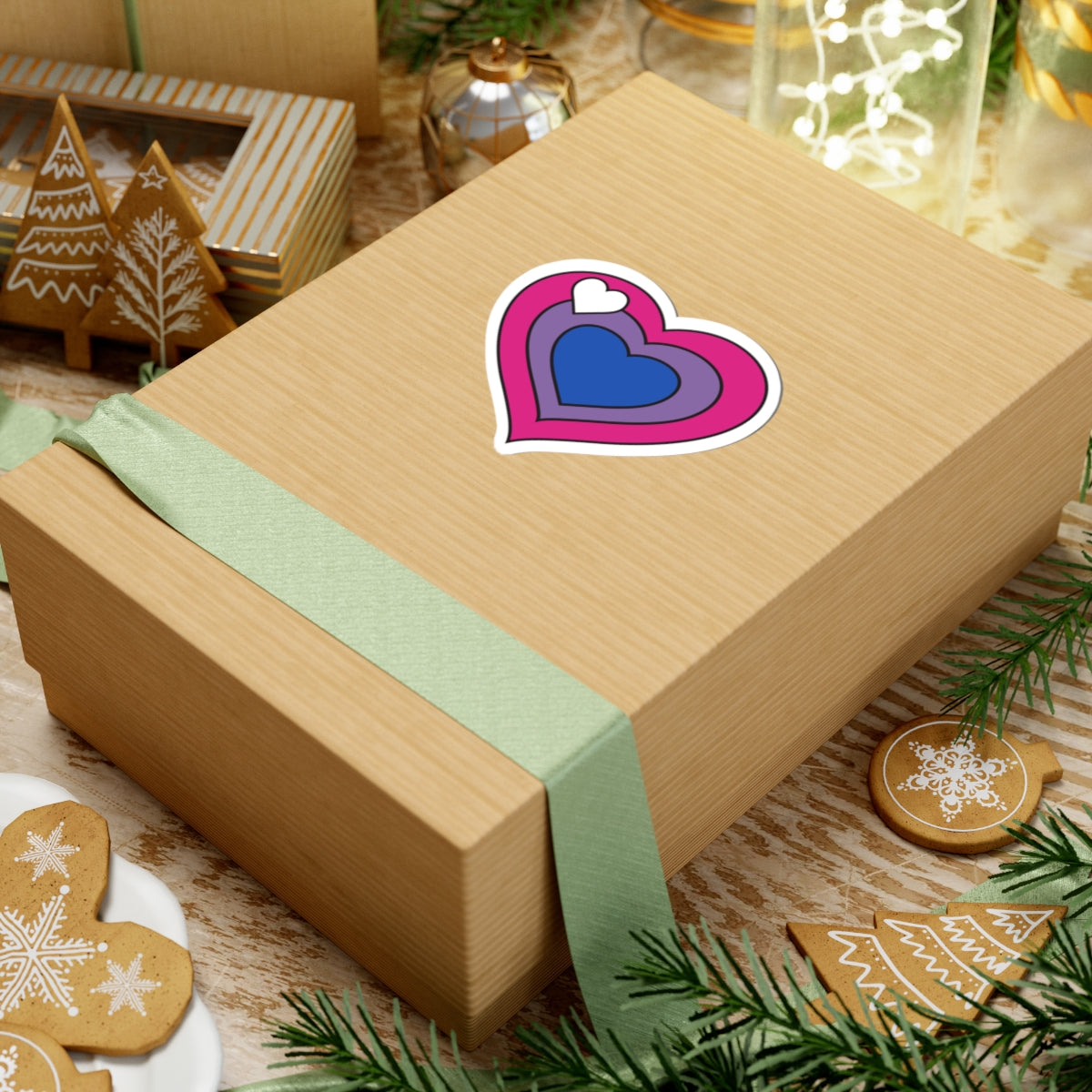 Gift box with a green ribbon on a festive background with cookies and Christmas decorations. Heart-shaped biromantic pride sticker centered on the box.