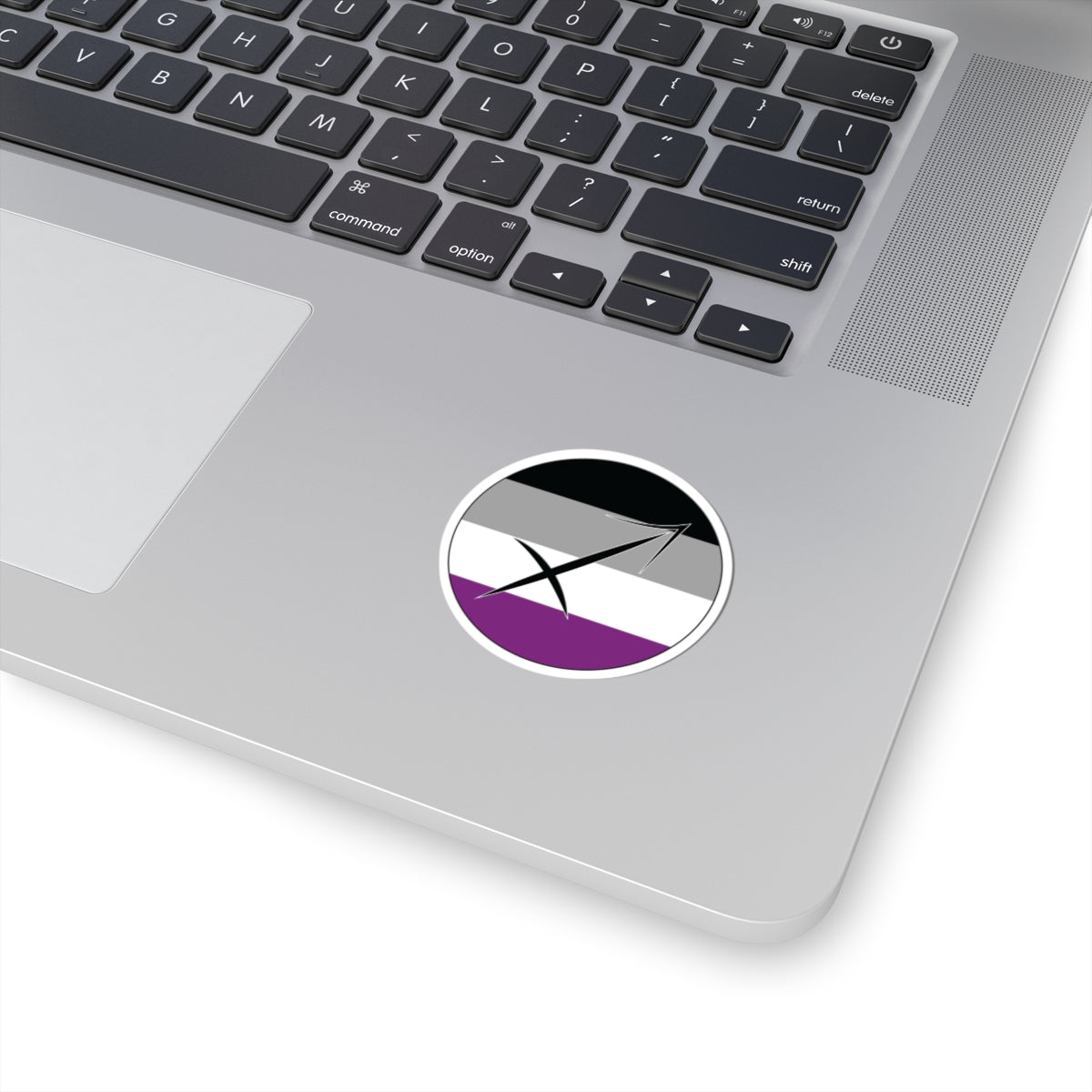 Close-up of the bottom right corner of a silver laptop featuring a 2 inch by 2 inch circular zodiac sticker with a centered Sagittarius glyph on an Asexual pride flag style background applied to the surface.