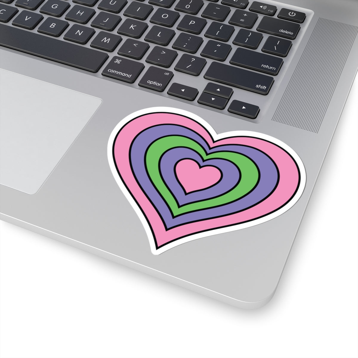 Close-up of the bottom right corner of a silver laptop featuring a 4 inch by 4 inch pride flag heart sticker in a Trigender flag style applied to the surface.