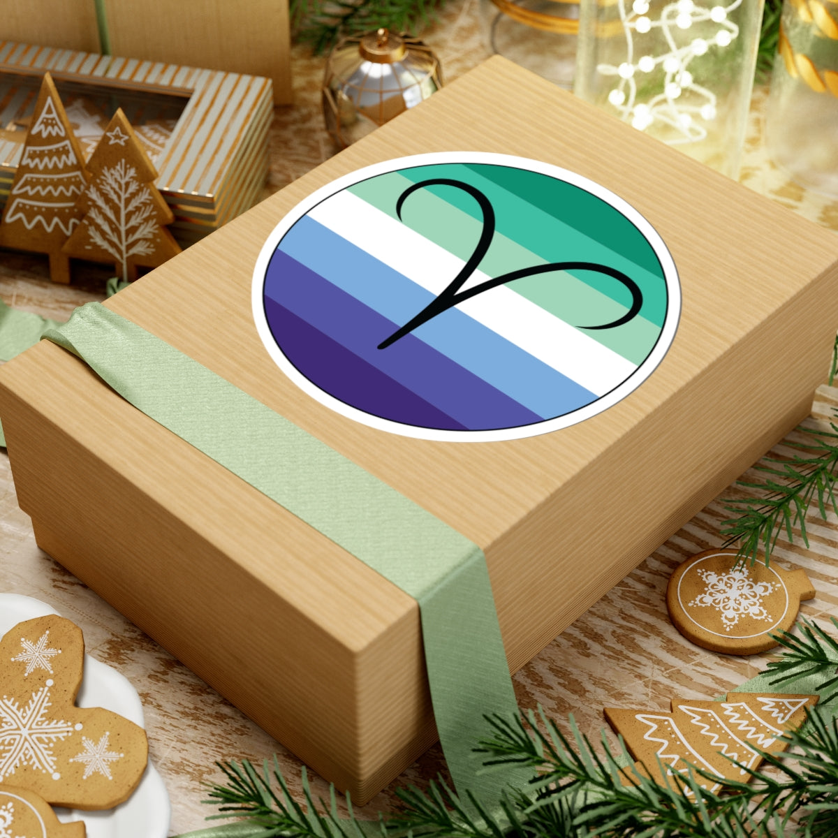 Gift box with a green ribbon on a festive background with cookies and Christmas decorations. Aires Gay Men pride sticker centered on the box.