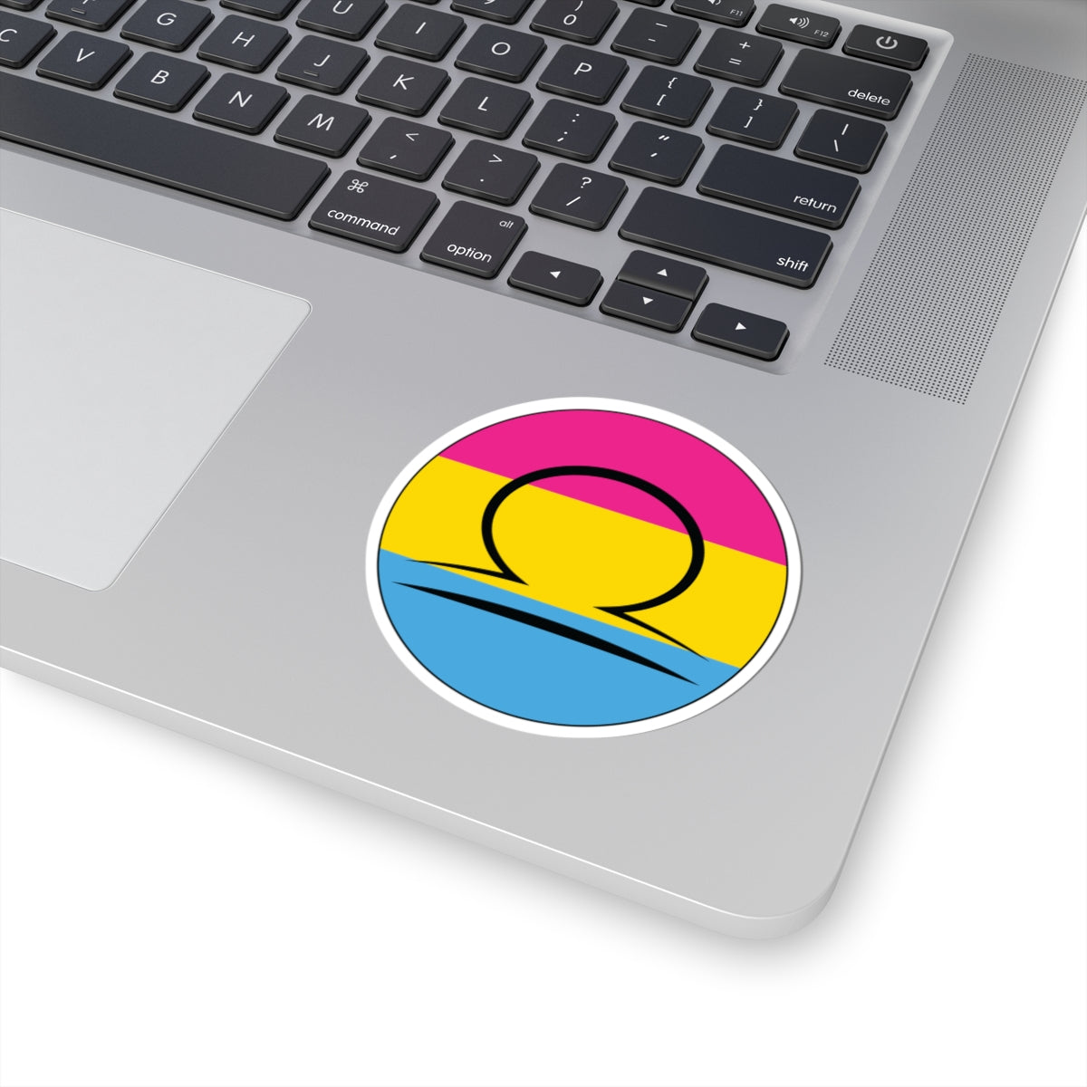 Close-up of the bottom right corner of a silver laptop featuring a 3 inch by 3 inch circular zodiac sticker with a centered Libra glyph on a Pansexual pride flag style background applied to the surface.