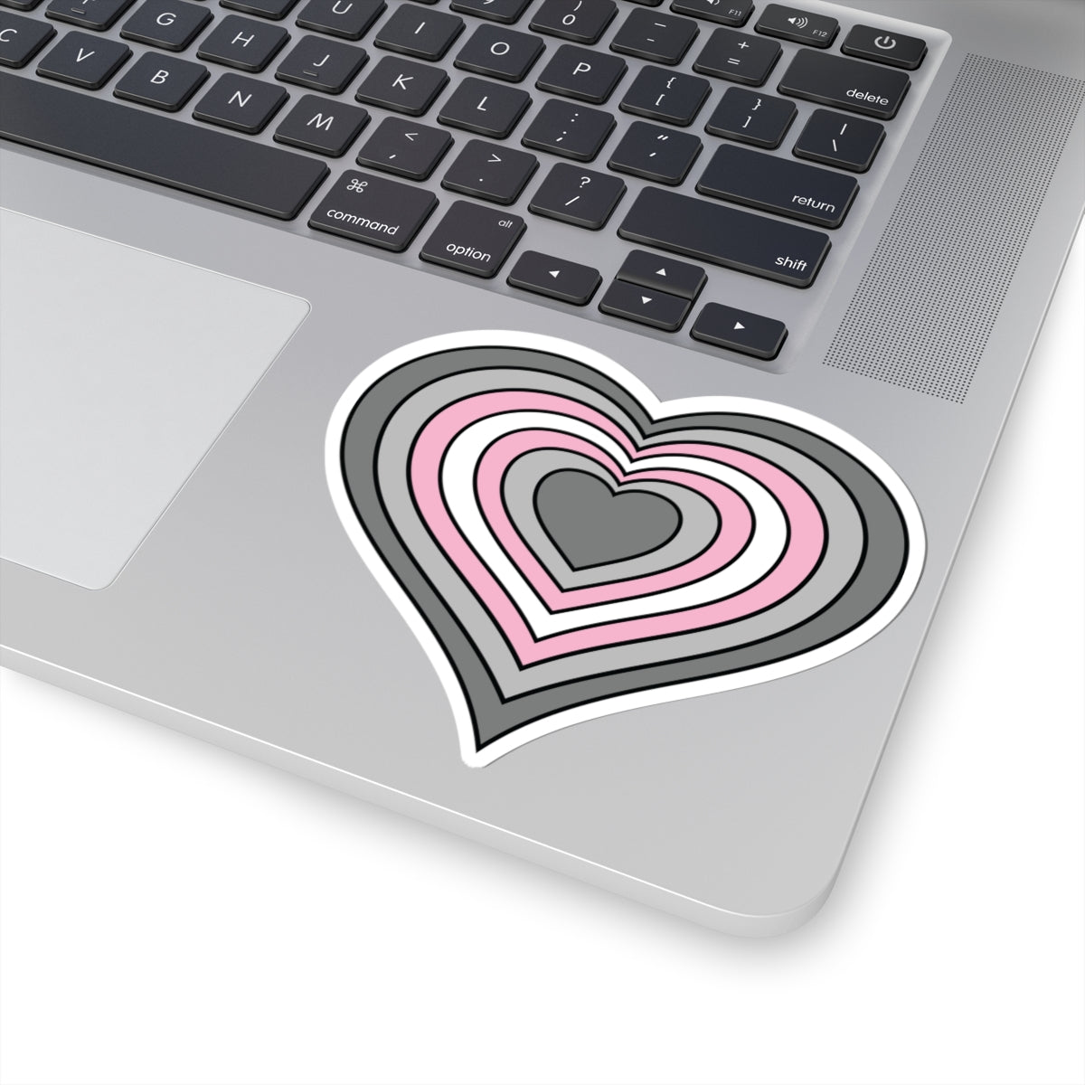 Close-up of the bottom right corner of a silver laptop featuring a 4 inch by 4 inch pride flag heart sticker in a Demigirl flag style applied to the surface.
