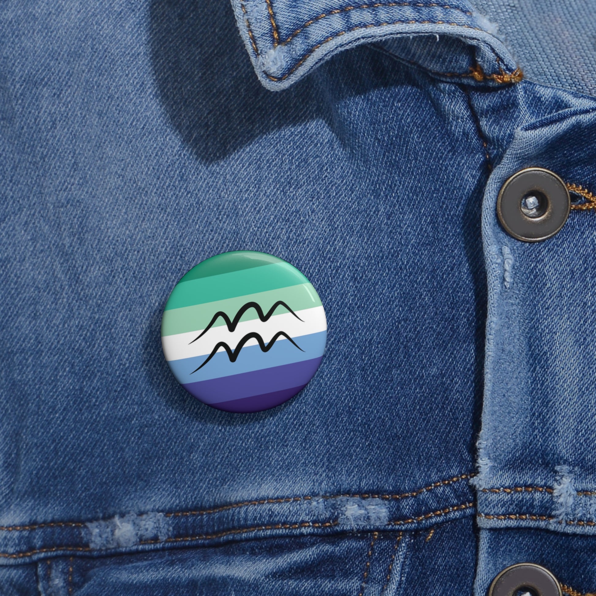 Close-up of a white circular pride button featuring the Aquarius zodiac glyph centered over a gay men (MLM) pride flag background in greens/teals, white, and blues, attached to a blue denim jacket at the right chest with the button clearly visible.