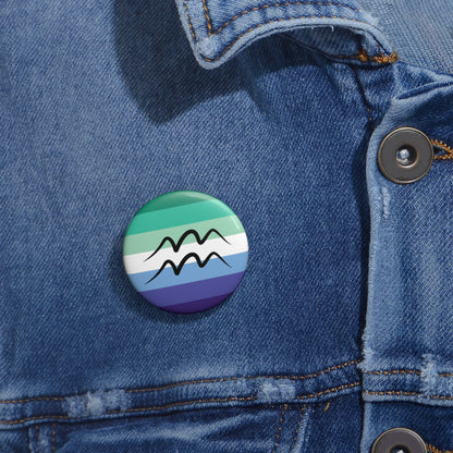 Close-up of a white circular pride button featuring the Aquarius zodiac glyph centered over a gay men (MLM) pride flag background in greens/teals, white, and blues, attached to a blue denim jacket at the right chest with the button clearly visible.