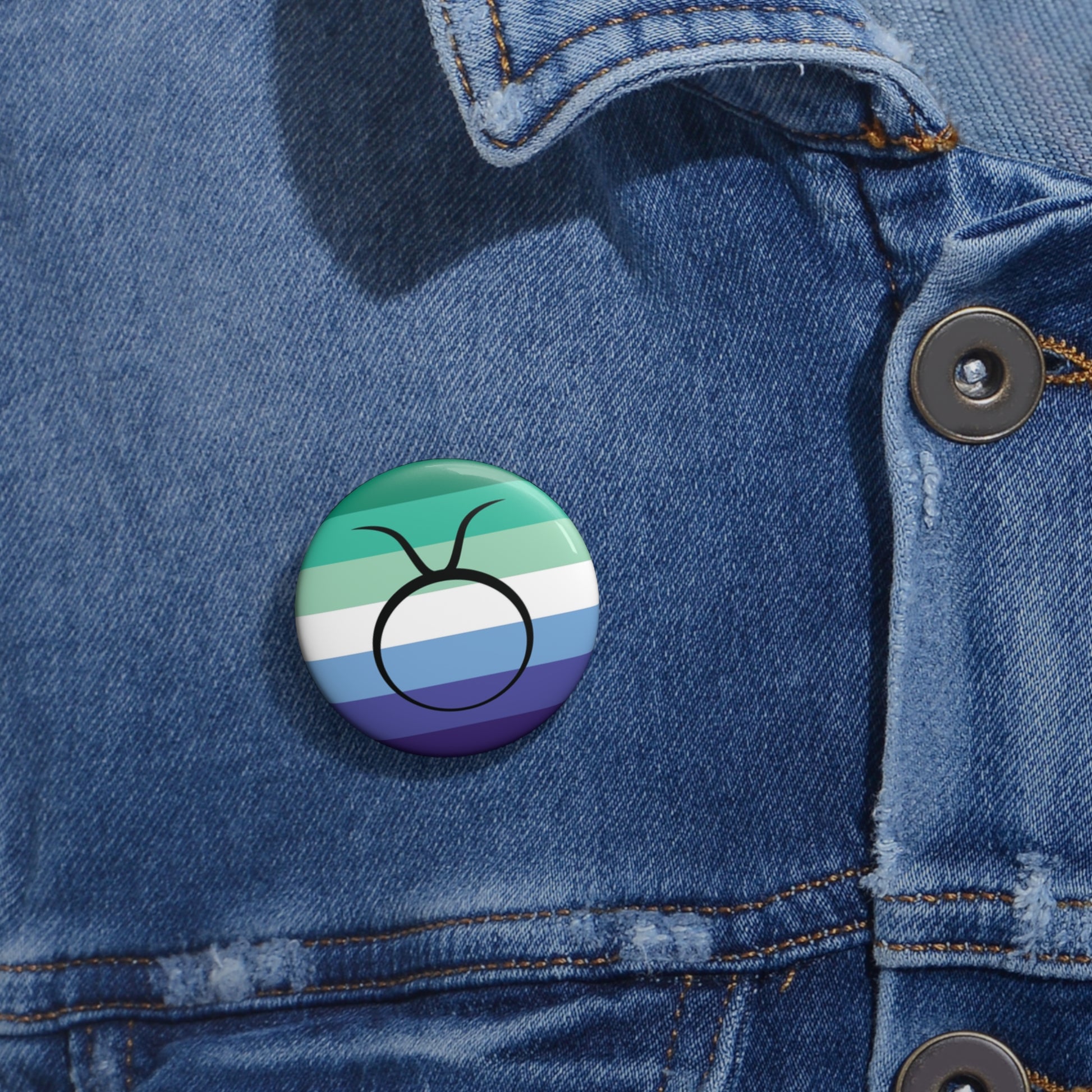 Close-up of a white circular pride button featuring the Taurus zodiac glyph centered over a gay men (MLM) pride flag background in greens/teals, white, and blues, attached to a blue denim jacket at the right chest with the button clearly visible.