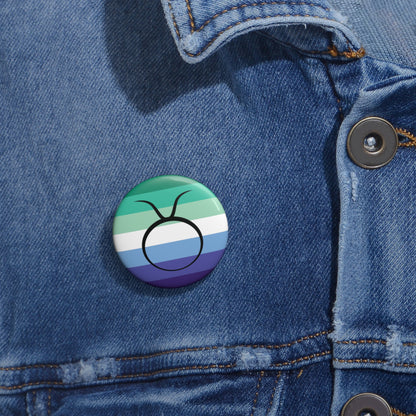 Close-up of a white circular pride button featuring the Taurus zodiac glyph centered over a gay men (MLM) pride flag background in greens/teals, white, and blues, attached to a blue denim jacket at the right chest with the button clearly visible.