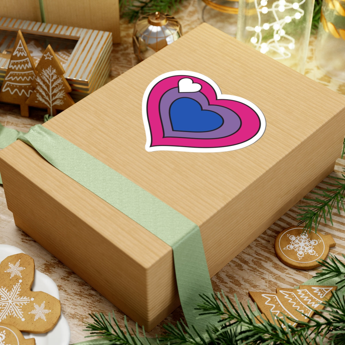Gift box with a green ribbon on a festive background with cookies and Christmas decorations. Heart-shaped biromantic pride sticker centered on the box.
