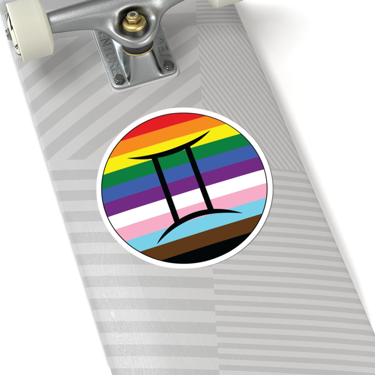 Close-up of the bottom of a silver skateboard featuring a 6 inch by 6 inch circular zodiac sticker with a centered Gemini glyph on a progress pride flag style background applied to the surface.