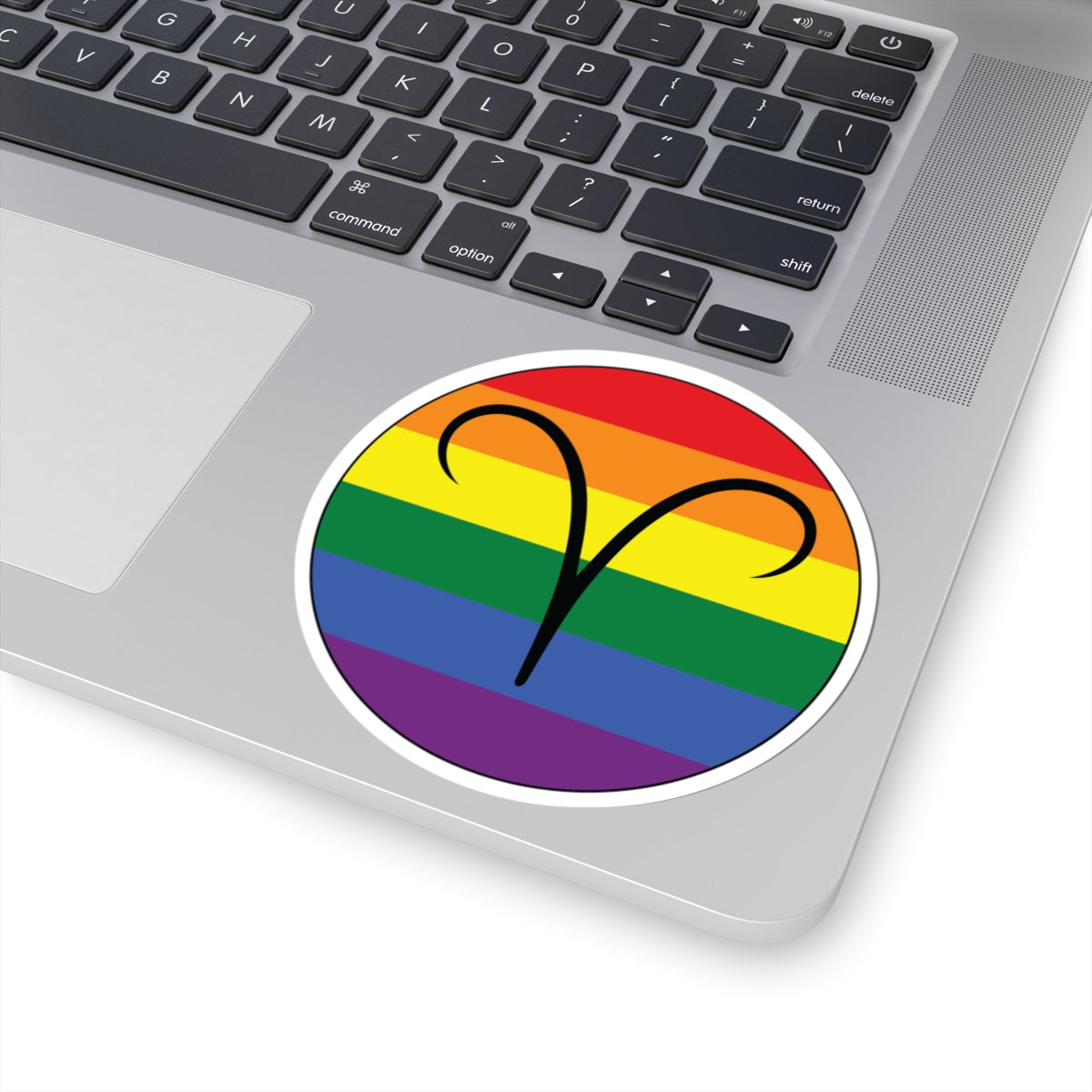 Close-up of the bottom right corner of a silver laptop featuring a 4 inch by 4 inch circular zodiac sticker with a centered Aires glyph on a Rainbow pride flag style background applied to the surface.