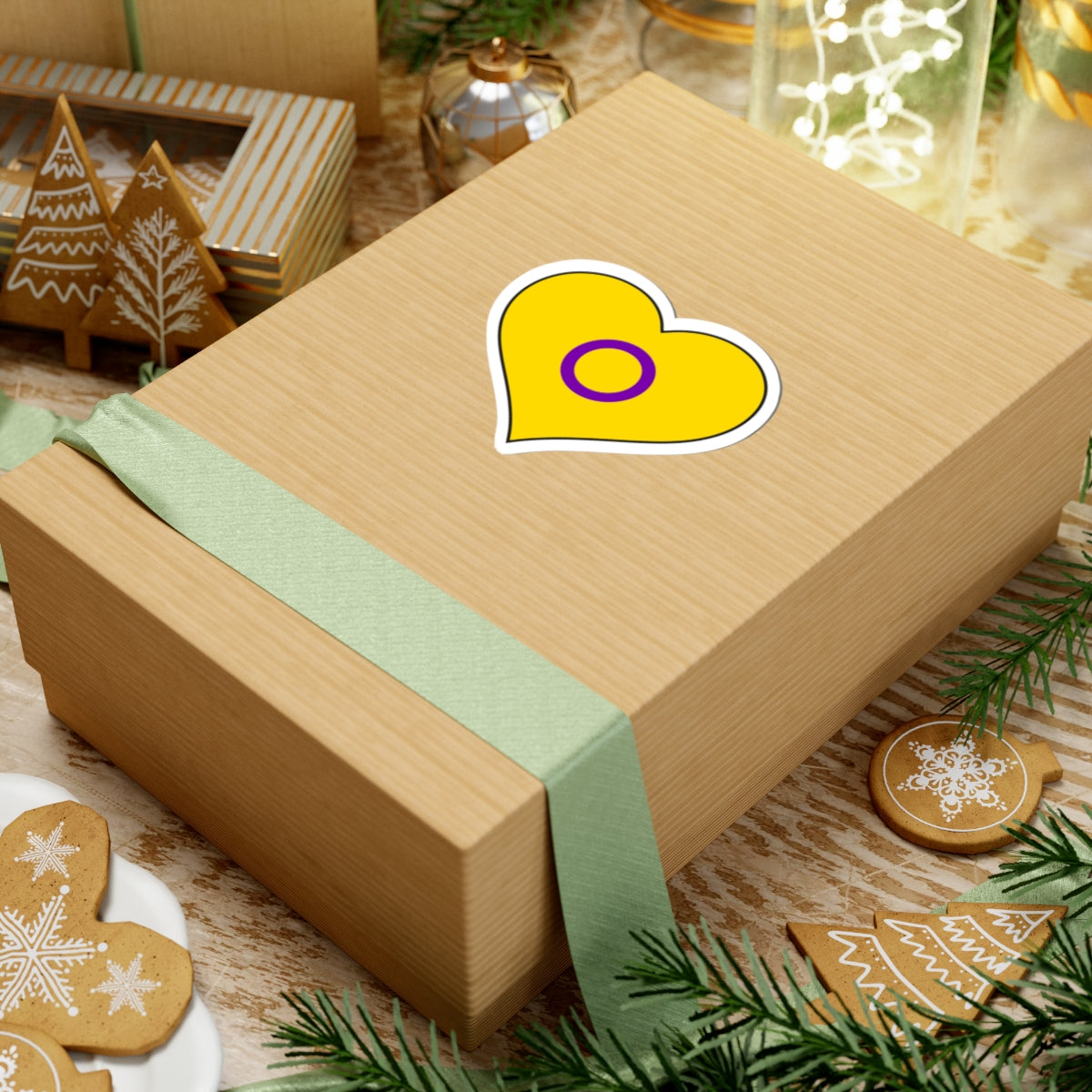 Gift box with a green ribbon on a festive background with cookies and Christmas decorations. Heart-shaped Intersex pride sticker centered on the box.
