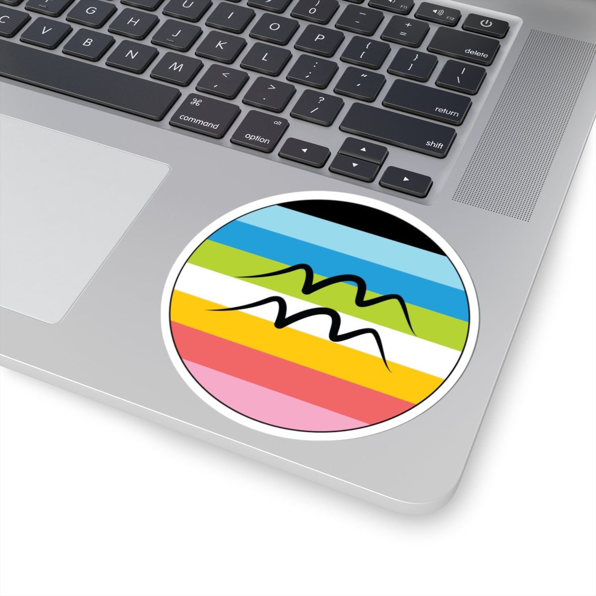 Close-up of the bottom right corner of a silver laptop featuring a 4 inch by 4 inch circular zodiac sticker with a centered Aquarius glyph on a Queer pride flag style background applied to the surface.