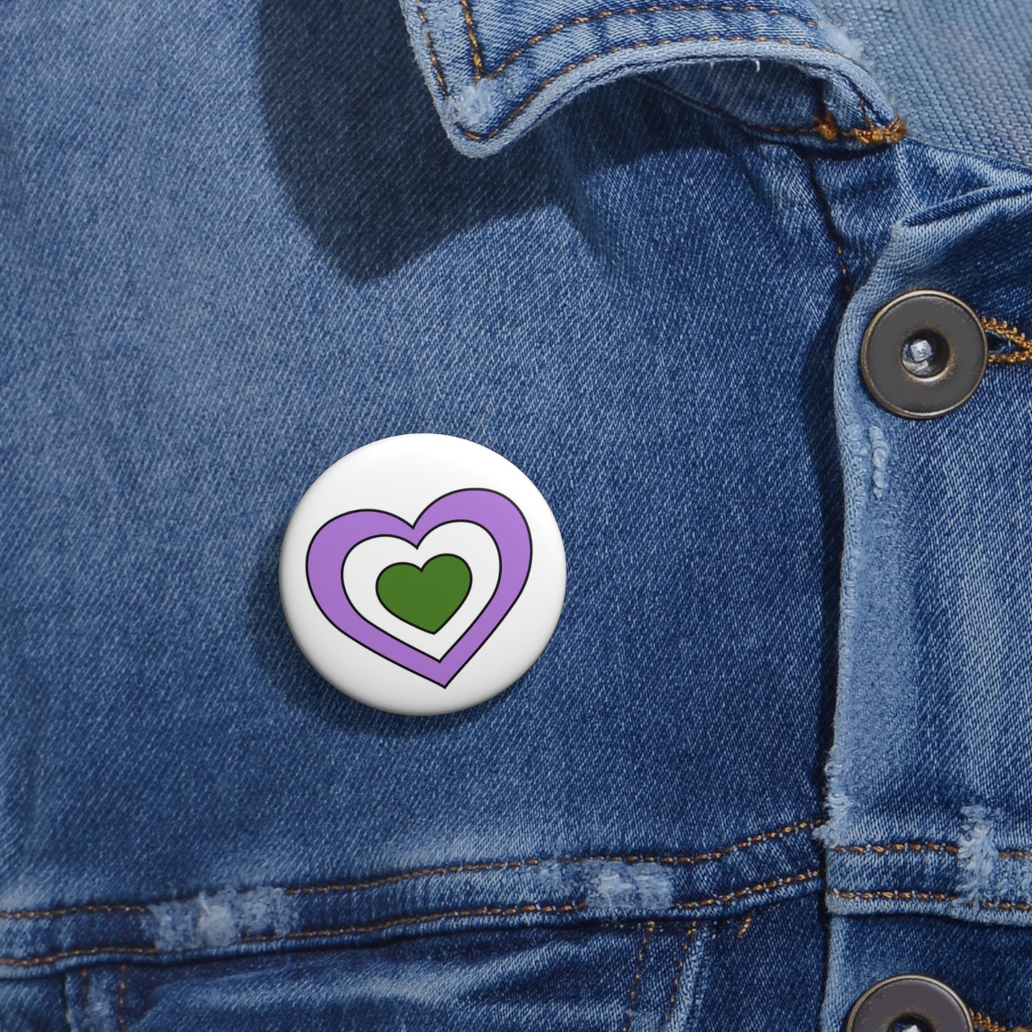 Close-up of a white circular pride button featuring a nested heart design inspired by the Genderqueer pride flag in purple, white and green, attached to a blue denim jacket at the right chest with the button clearly visible.