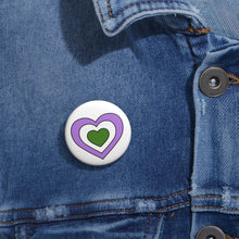 Close-up of a white circular pride button featuring a nested heart design inspired by the Genderqueer pride flag in purple, white and green, attached to a blue denim jacket at the right chest with the button clearly visible.