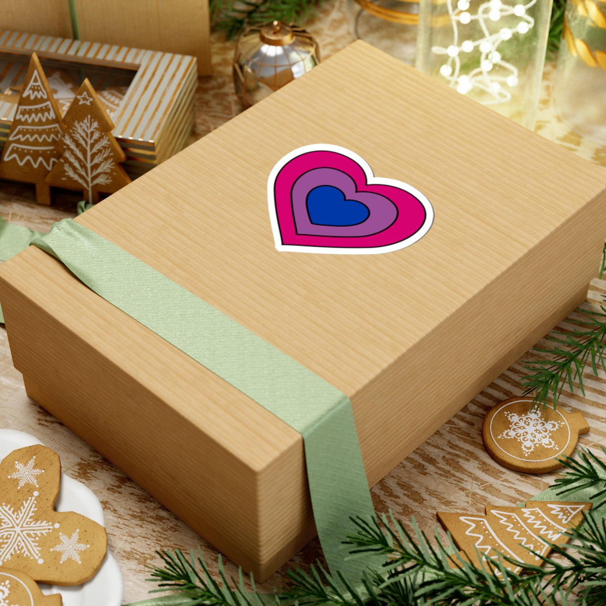Gift box with a green ribbon on a festive background with cookies and Christmas decorations. Heart-shaped Bisexual pride sticker centered on the box.