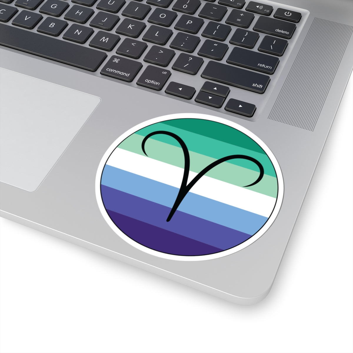 Close-up of the bottom right corner of a silver laptop featuring a 4 inch by 4 inch circular zodiac sticker with a centered Aires glyph on a Gay Men pride flag style background applied to the surface.