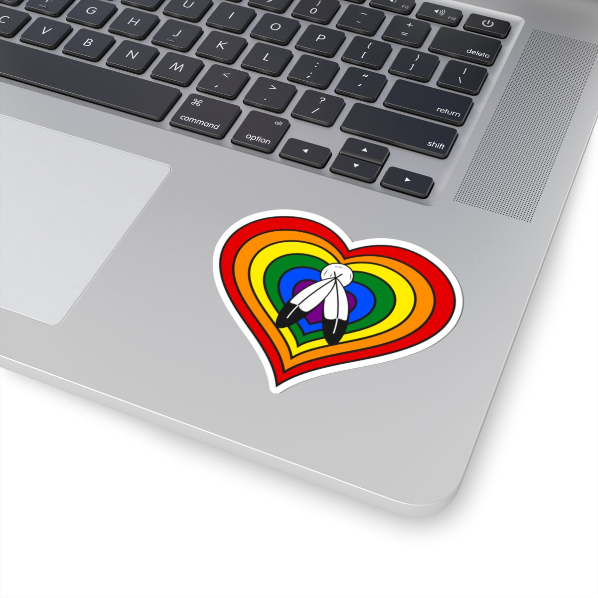 Close-up of the bottom right corner of a silver laptop featuring a 3 inch by 3 inch pride flag heart sticker in a Two-Spirit flag style applied to the surface.