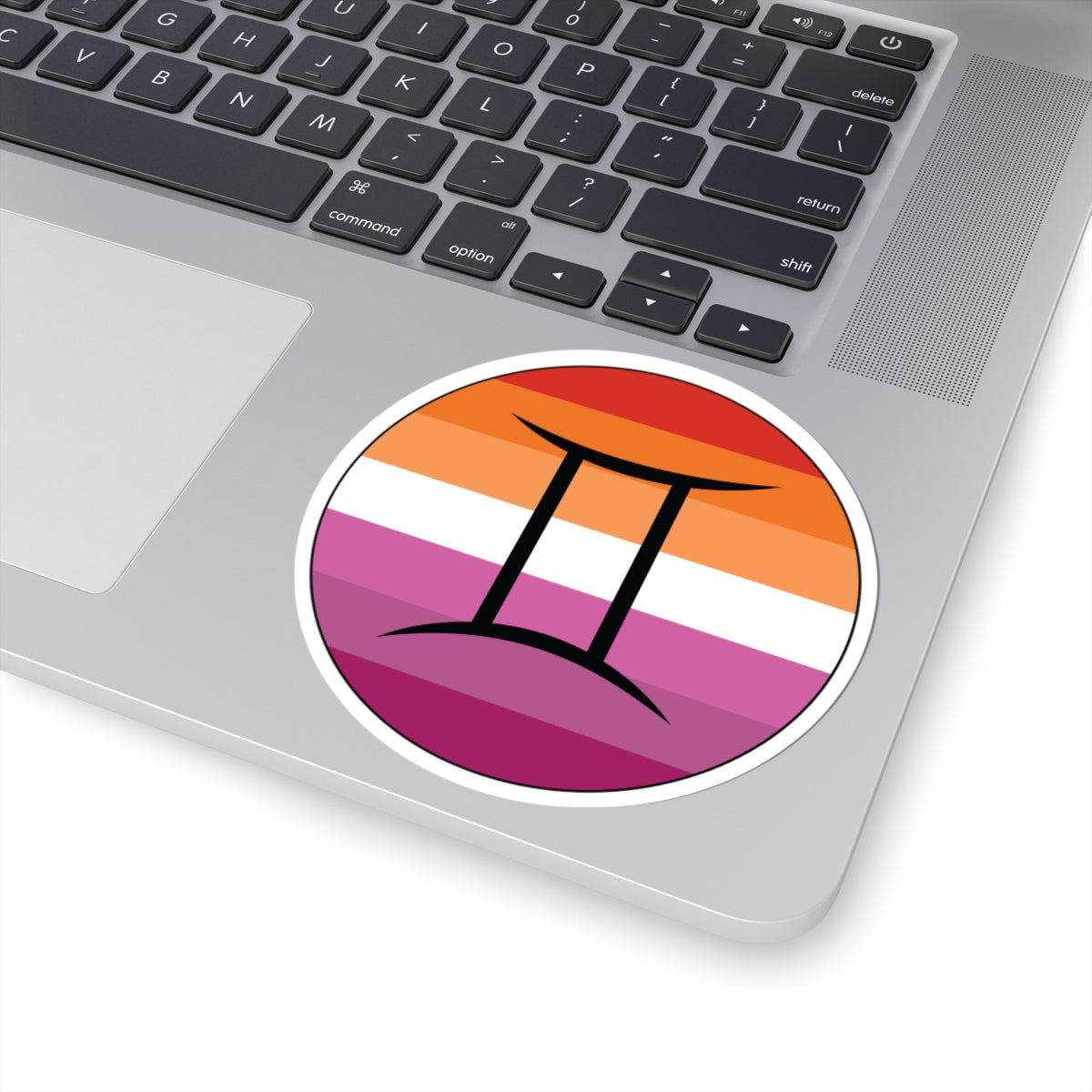 Close-up of the bottom right corner of a silver laptop featuring a 4 inch by 4 inch circular zodiac sticker with a centered Gemini glyph on a Lesbian pride flag style background applied to the surface.