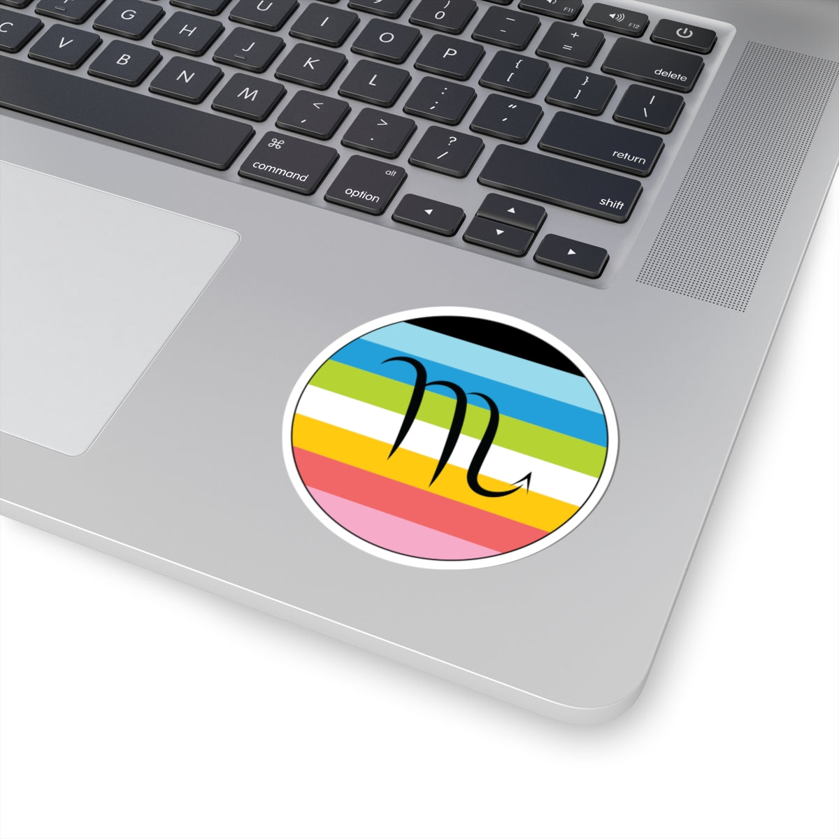 Close-up of the bottom right corner of a silver laptop featuring a 3 inch by 3 inch circular zodiac sticker with a centered Scorpio glyph on a Queer pride flag style background applied to the surface.