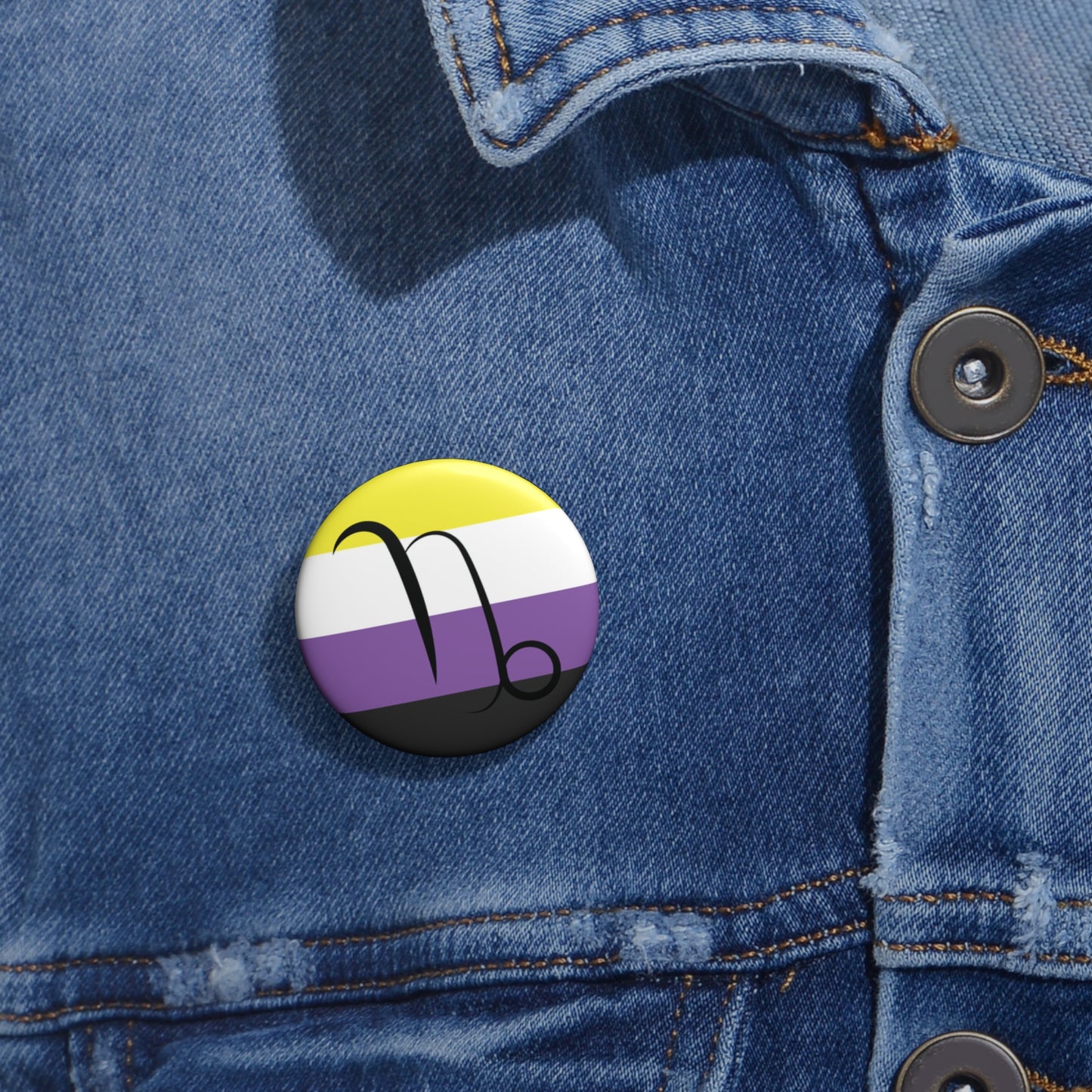 Close-up of a white circular pride button featuring the Capricorn zodiac glyph centered over a nonbinary pride flag background in yellow, white, purple, and black, attached to a blue denim jacket at the right chest with the button clearly visible.