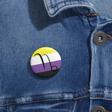 Close-up of a white circular pride button featuring the Capricorn zodiac glyph centered over a nonbinary pride flag background in yellow, white, purple, and black, attached to a blue denim jacket at the right chest with the button clearly visible.