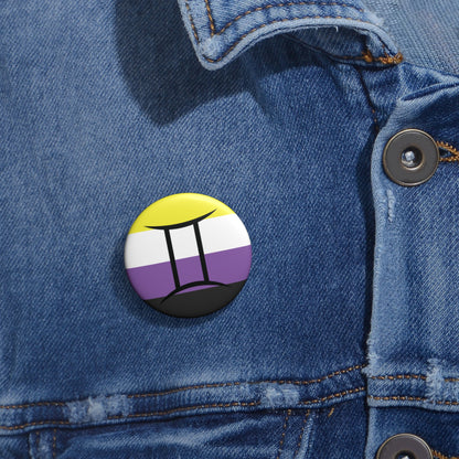 Close-up of a white circular pride button featuring the Gemini zodiac glyph centered over a nonbinary pride flag background in yellow, white, purple, and black, attached to a blue denim jacket at the right chest with the button clearly visible.