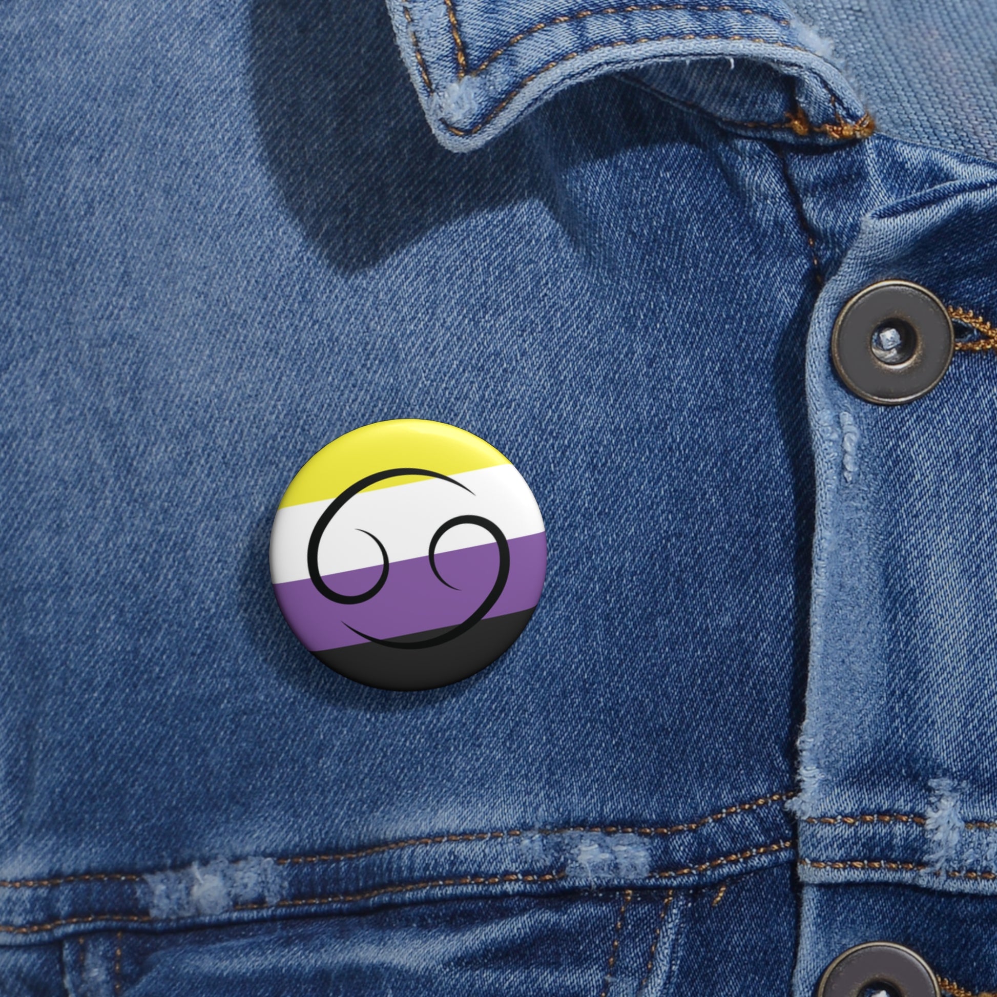 Close-up of a white circular pride button featuring the Cancer zodiac glyph centered over a nonbinary pride flag background in yellow, white, purple, and black, attached to a blue denim jacket at the right chest with the button clearly visible.