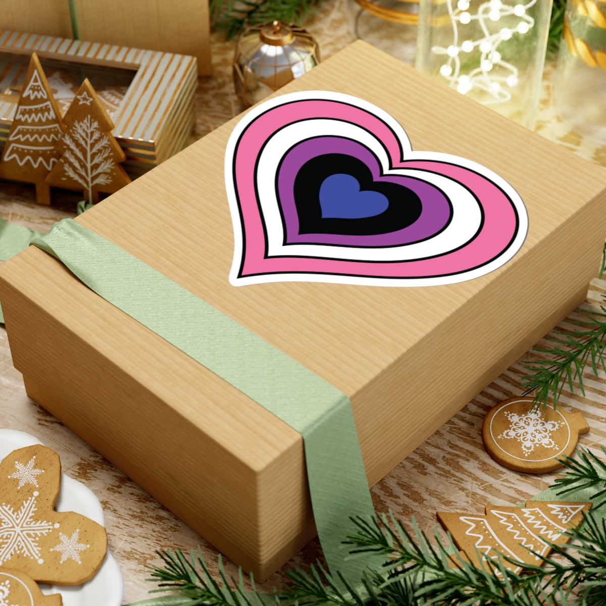 Gift box with a green ribbon on a festive background with cookies and Christmas decorations. Heart-shaped Genderfluid pride sticker centered on the box.