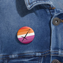 Close-up of a white circular pride button featuring the Sagittarius zodiac glyph centered over a lesbian pride flag background in oranges, white, and pinks attached to a blue denim jacket at the right chest with the button clearly visible.