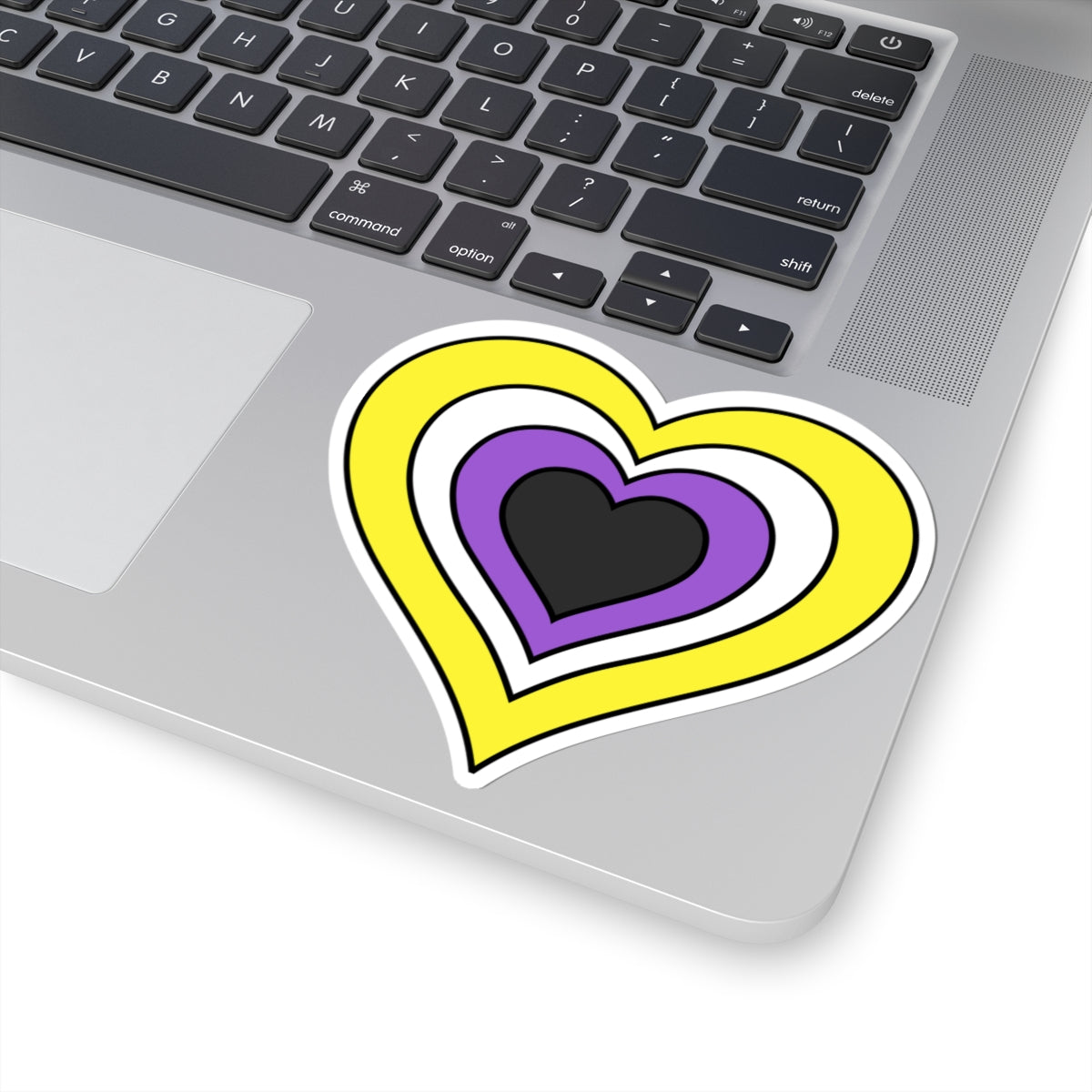 Close-up of the bottom right corner of a silver laptop featuring a 4 inch by 4 inch pride flag heart sticker in a Nonbinary flag style applied to the surface.