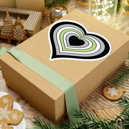 Gift box with a green ribbon on a festive background with cookies and Christmas decorations. Heart-shaped Agender pride sticker centered on the box.