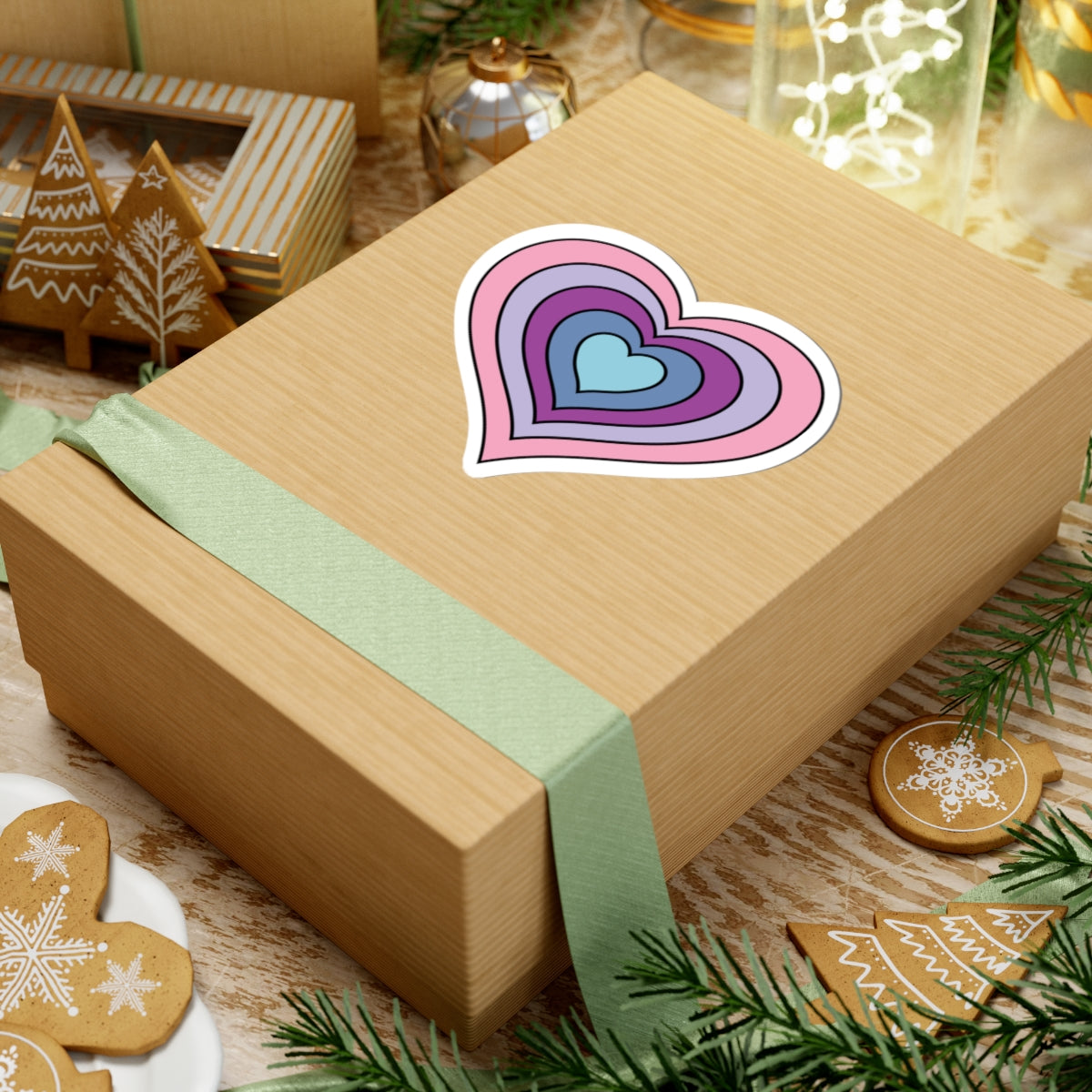 Gift box with a green ribbon on a festive background with cookies and Christmas decorations. Heart-shaped Omnigender pride sticker centered on the box.