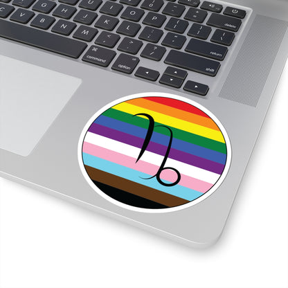 Close-up of the bottom right corner of a silver laptop featuring a 4 inch by 4 inch circular zodiac sticker with a centered Capricorn glyph on a progress pride flag style background applied to the surface.